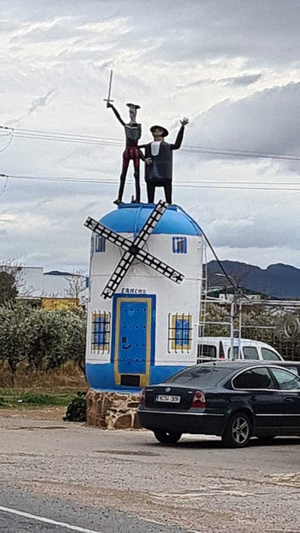 Kleines Haus in Form einer weißen Windmühle mit blauen Türen und Fenstern, gekrönt von Don Quijote und Sancho Panza als Figuren – skurriles Wahrzeichen am Straßenrand – Wohnmobil-Rundreise Spanien
